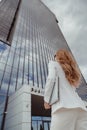 Young business woman in white suit stands at enterance of skyscraper with laptop. Bottom view