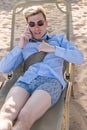 Young businessman downshifter talks on the phone lying on a sun lounger on the beach in a shirt, tie and boxer shorts