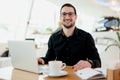 Young businessman in eyeglasses drinking coffee while working on laptop computer in coffeehouse, male freelancer writing Royalty Free Stock Photo