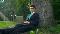 Young businessman with laptop sitting under tree, working remotely in park Royalty Free Stock Photo