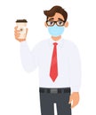 Young businessman in safety medical mask showing coffee cup. Person holding paper mug. Male character in formal wear