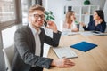 Young businessman sit at table in meeting room. He talk on phone and smile to camera. Two young women sit behind and