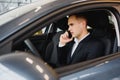 Young businessman sits in luxury car and talks on phone. He looks straight forward. Guy drives car. He holds one hand on steering