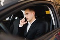 Young businessman sits in luxury car and talks on phone. He looks straight forward. Guy drives car. He holds one hand on steering