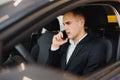 Young businessman sits in luxury car and talks on phone. He looks straight forward. Guy drives car. He holds one hand on steering