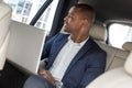 Young businessman sitting on back seat in car working on laptop looking out the window thoughtful