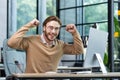 A young businessman, student, freelancer, programmer sits in the office at the table and celebrates success, victory