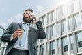 Young businessman in suit and tie is standing outdoor, drinking coffee and talking on his cell phone. Royalty Free Stock Photo