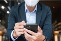 Young Businessman in suit wearing surgical face mask and using smartphone, man typing touchscreen mobile phone in office or cafe. Royalty Free Stock Photo