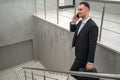Young businessman with suitcase in stylish suit talking by phone while walking by stairs in modern business building Royalty Free Stock Photo