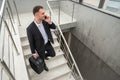 Young businessman with suitcase in stylish suit talking by phone while walking by stairs in modern business building Royalty Free Stock Photo