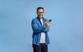 Young businessman texting on smart phone and looking at camera while standing on blue background