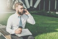 Young businessman in white shirt and tie is sitting outside on bench, drinking coffee and talking on his cell phone Royalty Free Stock Photo