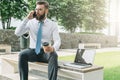 Young businessman in white shirt and tie is sitting outside on bench,drinking coffee and talking on his cell phone. Royalty Free Stock Photo