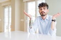 Young businesss man sitting on white table clueless and confused expression with arms and hands raised