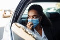 Young businesswoman being a taxi passenger and having a ride wearing and adjusting medical mask for health protection. Business
