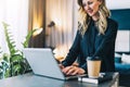 Young businesswoman in black blouse is standing indoor, working on computer. Girl freelancer, entrepreneur works at home