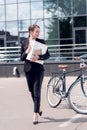 young businesswoman in earphones with documents walking on street with bicycle