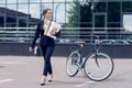 young businesswoman in earphones with documents walking on street with bicycle