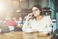 Young businesswoman in glasses and white sweater is sitting in cafe at wooden table and talking on cell phone. Royalty Free Stock Photo