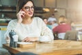 Young businesswoman in glasses and white sweater is sitting in cafe at wooden table and talking on cell phone Royalty Free Stock Photo
