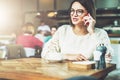 Young businesswoman in glasses and white sweater is sitting in cafe at wooden table and talking on cell phone Royalty Free Stock Photo