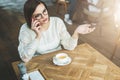 Young businesswoman in glasses and white sweater is sitting in cafe at wooden table and talking on cell phone. Royalty Free Stock Photo