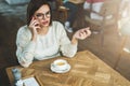 Young businesswoman in glasses and white sweater is sitting in cafe at wooden table and talking on mobile phone Royalty Free Stock Photo