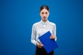 Young businesswoman seriously looking into the camera while holding a blue clipboard and a pen, on blue background