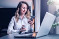 Young businesswoman in shirt is sitting in office at table in front of computer, using smartphone, looks at phone screen