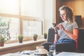 Young businesswoman sits on sofa near window,uses smartphone. Girl chatting, blogging, checking email, working. Royalty Free Stock Photo