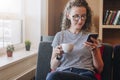 Young businesswoman sits on sofa,uses smartphone,working, drinking coffee.Hipster girl is chatting,blogging,checking Royalty Free Stock Photo