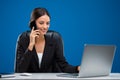 Young businesswoman sitting at her desk listening carefully to her phone and typing at the computer, isolated on blue background Royalty Free Stock Photo