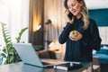 Young businesswoman is standing indoor near table in front of computer, while talking on cell phone and holding apple. Royalty Free Stock Photo