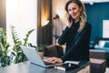 Young businesswoman standing indoor, working on computer, while talking on cell phone. Girl entrepreneur works at home.