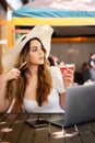 Young businesswoman wearing big hat working at the computer in cafe. Attractive girl downshifter working at a laptop in