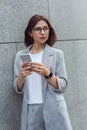 Business casual. Young lady in eyeglasses standing isolated on wall using app on smartphone looking aside curious close