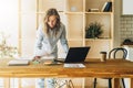 Young businesswoman woman is standing near kitchen table,reading documents,uses laptop,working, studying. Royalty Free Stock Photo