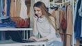 Young businesswoman is working with laptop and talking on mobile phone in her shop. Clothes and customer in background Royalty Free Stock Photo