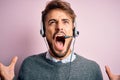 Young call center agent man with beard wearing headset over isolated pink background crazy and mad shouting and yelling with Royalty Free Stock Photo