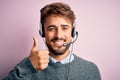 Young call center agent man with beard wearing headset over isolated pink background doing happy thumbs up gesture with hand Royalty Free Stock Photo