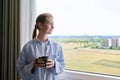 Young calm dreaming woman with cup in her hands, home window background