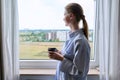 Young calm dreaming woman with cup in her hands, home window background