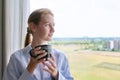 Young calm dreaming woman with cup in her hands, home window background