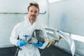 Young car paint service worker stands in a automobile painting booth with a gun and a protective mask in his hands. Royalty Free Stock Photo