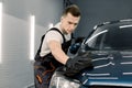 Young car wash worker man, wearing protective clothes and gloves, cleaning modern dark blue car with microfiber cloth Royalty Free Stock Photo