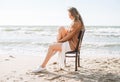 Young carefree beautiful woman with long hair in sweater sitting on chair on sea beach Royalty Free Stock Photo