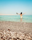 Young carefree Caucasian woman tourist dance on the beach in turquoise water alone with hands up hold slippers on summer vacation