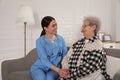 Young caregiver talking to senior woman on sofa in room. Home health care service Royalty Free Stock Photo