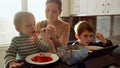 Young caring mother feeding her two sons with soup on the kitchen filled with sunset light